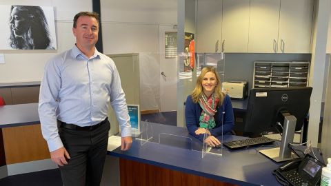 A man stands in front of a desk while a women sits behind.