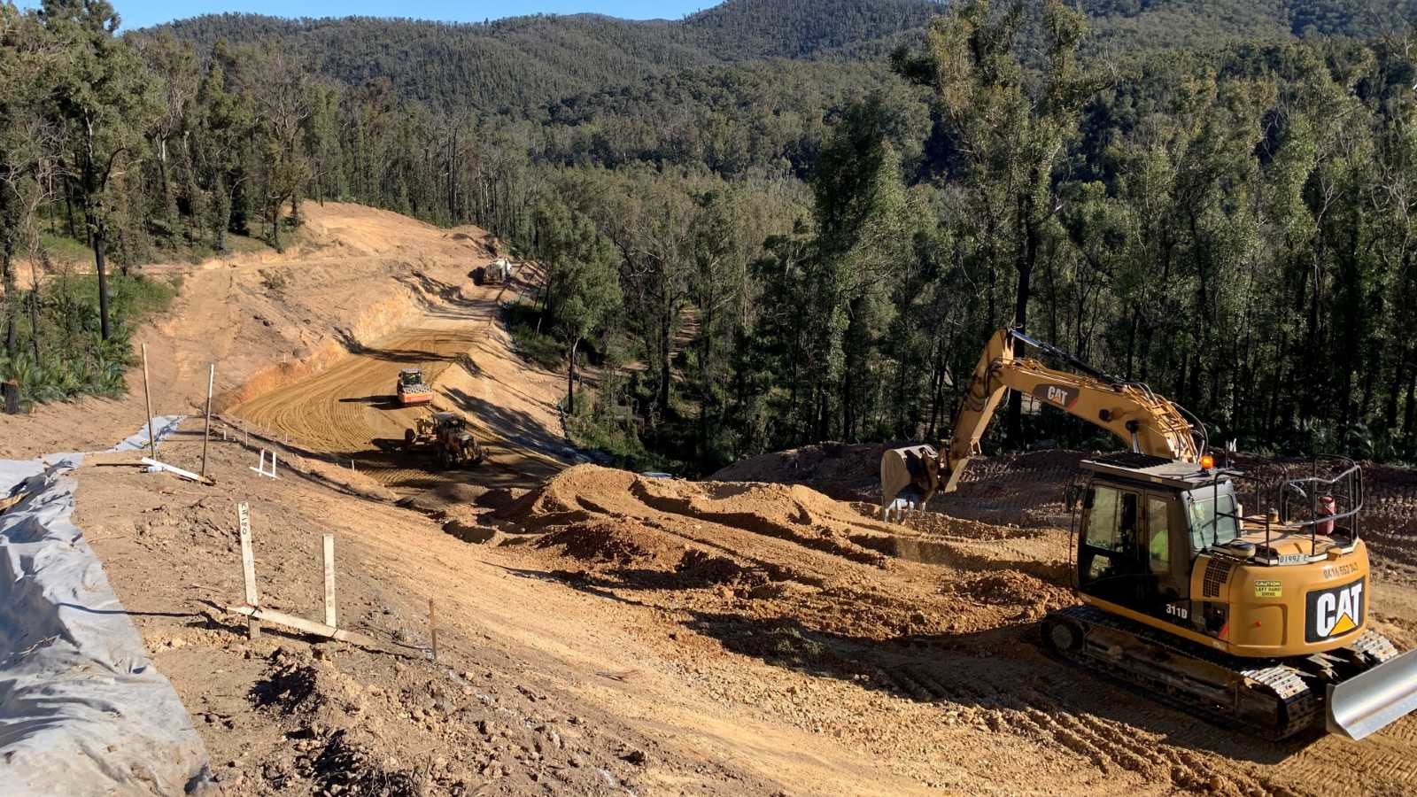 A large excavator works to clear a road through mountainous forest.