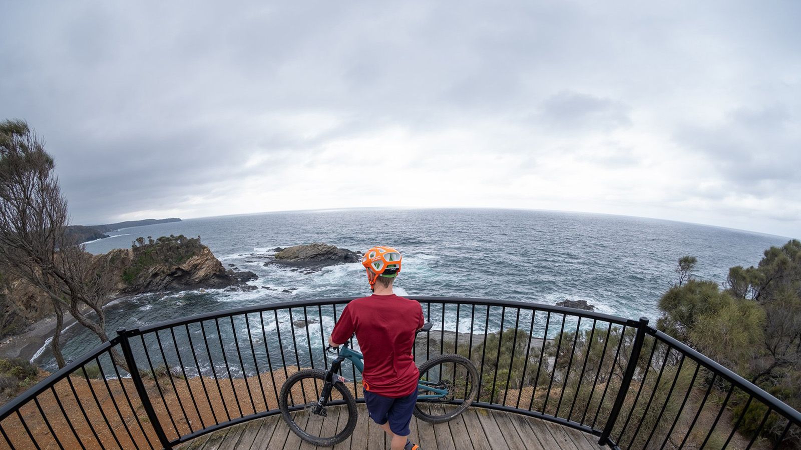 A cyclist standing next to his bike looking out over the water at a lookout with a railing banner image