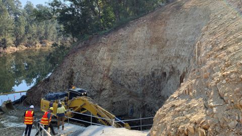 Three workers stand alongside an excavator, which is digging a hole at the base of a hill, next to a river.