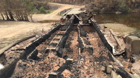Burnt out bridge across large sandy river bed.