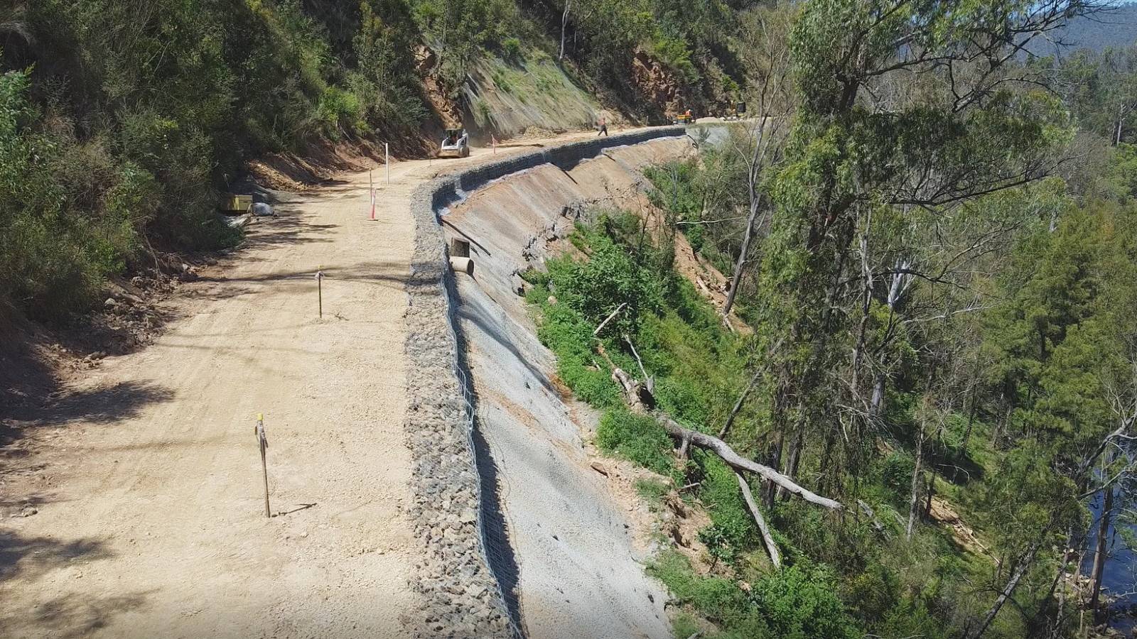 A dirt road is supported by a wall of gabion baskets and shotcrete on the steep downhill slope in a forest.