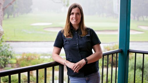 Dark haired woman wearing blue shirt leaning on railing with gold course in background.