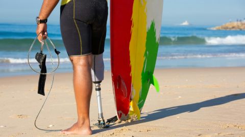 Half body image of a male surfer with a prosthetic leg, holding a surfboard next to the ocean.